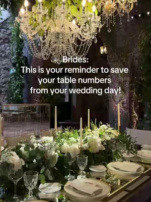 A wedding reception table is richly decorated and set at a formal event. A sparkling chandelier provides overhead lighting, and the table is adorned with white flowers, candles, and ornate glassware. The setting suggests sophistication and elegance.