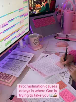 An indoor desk setup with a computer, laptop, and papers, with a pink aesthetic. A visible text overlay at the bottom reads: "Procrastination causes delays in where God is trying to take you".