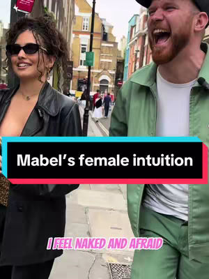 A candid shot of two people in an urban setting. A woman on the left is wearing sunglasses and a black leather jacket, smiling broadly. The right side of the image features a man in a green outfit, laughing. Overlaid are text elements on the image with a street and building background.