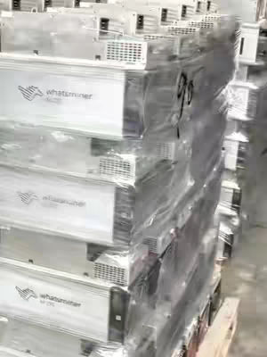 A stack of wrapped Bitcoin mining machines. The machines are silver and have the "whatsminer" logo visible. They are wrapped in clear plastic and stacked on a wooden palette. The background is a blurry industrial setting.