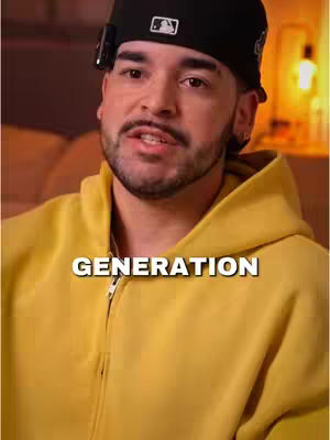 A man wearing a black baseball cap and a yellow hoodie, with the text 'GENERATION' overlaid. He has a mustache and a small beard, and appears to be speaking. The background is slightly blurred.