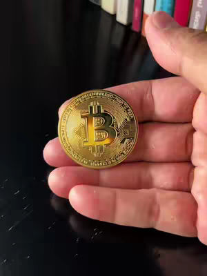 A hand holds a golden Bitcoin coin against a dark background with a row of books in the upper right corner.