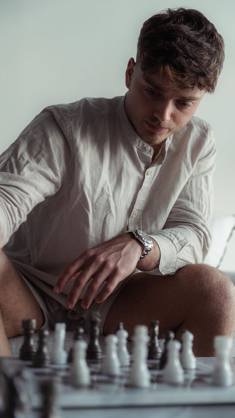 A medium shot of a man in a light-colored shirt engrossed in a game of chess. He sits with a focused expression, his hand resting on the table near the chess pieces, in a low angle shot emphasizing his face and watch.