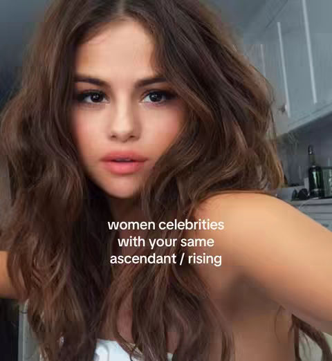 A selfie-style image featuring a woman with long brown wavy hair. The woman is looking directly at the camera with a slight smile, her lips are a light pink. The background is blurred but appears to be a kitchen. Text overlayed at the bottom reads 'women celebrities with your same ascendant / rising'.