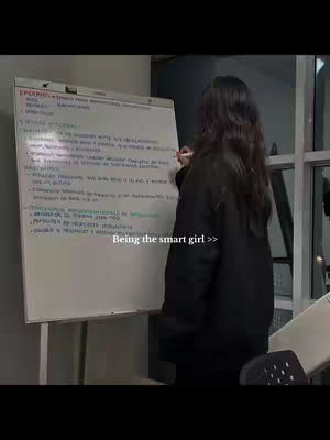 A woman with long dark hair, seen from the back, is standing in front of a whiteboard, writing on it with a pink marker. The whiteboard is filled with handwritten notes. The background is blurred, suggesting an indoor setting. Text overlay at the bottom says 'Being the smart girl >>'.