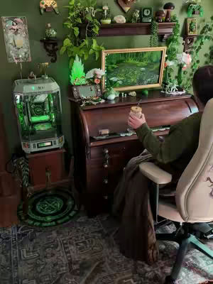 A cozy room setup with a gaming PC and desk decorated with plants and aesthetic items. A person is sitting in a chair, holding a warm beverage, with a painting on a desk in front of them.