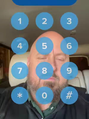 A man's face dominates the image, overlaid with blue circles containing numbers and symbols resembling a phone keypad. The man has a bald head and a full beard, and his eyes are closed. The background suggests the inside of a vehicle.