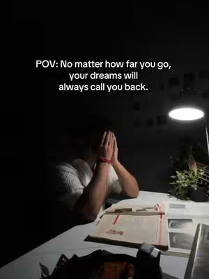 The image presents a dimly lit scene. A person is seated at a desk, their face obscured by their hands, suggesting introspection or stress. A desk lamp illuminates the area, highlighting books, and a small plant. The composition is simple with dark tones, with focused lighting directing attention.