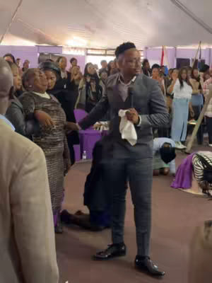The image is taken indoors, presumably in a church or event venue. In the center, a man wearing a dark gray suit and holding a white cloth is looking towards the right, appearing to address a crowd. To his left, a woman in a patterned dress is also looking in the same direction, with her hands near her chest. The background reveals a large gathering of people, some looking towards the speakers and some kneeling, suggesting a religious service or gathering. The color palette leans towards neutral tones with the subjects and background items.