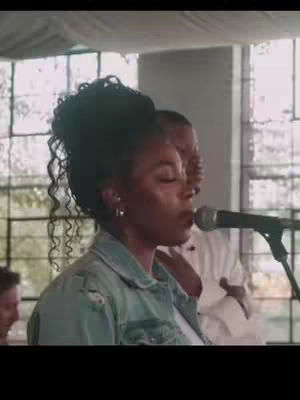 A woman with curly hair wearing a denim jacket is singing into a microphone mid-performance. Behind her are other individuals, and a window with a fabric draped over the top adds a softer touch to the visual.