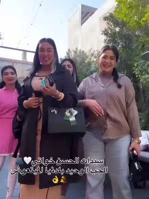 A group of stylish women pose together outdoors, smiling. The woman in the center has long dark hair and holds a black bag with a gift. They are standing in front of a building with trees and what looks like a patio. The women are wearing a variety of outfits, including a pink dress, a patterned blouse, and a long black coat over a top.