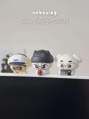 The image captures three ENHYPEN character figurines arranged on a white shelf against a simple neutral background. The figurines are depicted with cartoon-like features, each sporting unique accessories such as hats, glasses, and props, including a camera and a small item held in the figurines' hands. A graphic text overlay at the top reads 'unboxing EN-hippers,' and the overall composition has an inviting, collectible-focused appearance.