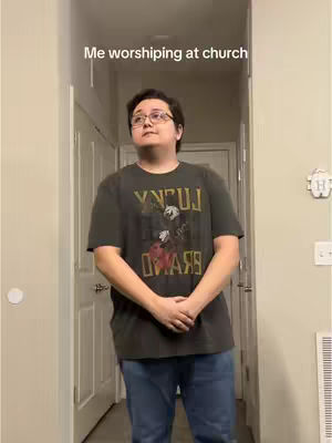 A person with glasses, hands clasped, staring upwards in what appears to be a hallway. Overlaid text reads 'Me worshiping at church'. The person is wearing a t-shirt with a design.