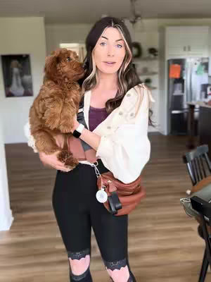 A woman stands holding a small, fluffy brown dog in an indoor setting. She wears a white jacket, black leggings, and a brown bag across her chest. The background shows a domestic interior with wooden flooring and a partially visible kitchen.