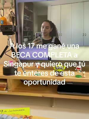 A woman is holding a cell phone in front of a mirror, taking a selfie. Text overlays the image in multiple lines, with the first line reading, "A los 17 me gané una," and the following lines include "BECA COMPLETA a Singapur y quiero que tú te enteres de esta oportunidad". There is a desk with many items.