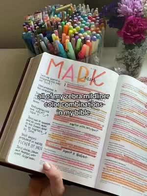A top-down shot shows an open Bible with text highlighted in various colored pens, alongside a jar of multicolored pens and a bouquet of flowers, all sitting on a white table.