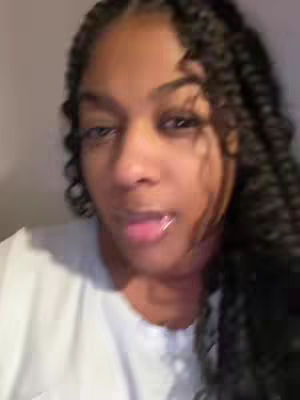 A close-up shot of a woman with dark, braided hair. She is looking slightly off-camera with a neutral expression and her tongue is sticking out. She is wearing a white top, and the background is a solid, neutral color.