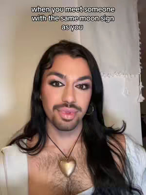 A person with long dark hair, makeup, and a mustache is the focal point. Above the person is some text that sets the scene. The person is wearing a white top and a heart-shaped pendant.