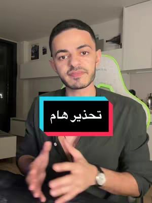 A man is centered in the shot, with a teal and red text overlay across his chest. He is in front of a wall, and his hands gesture towards the camera. Behind him are some shelves with items on them.