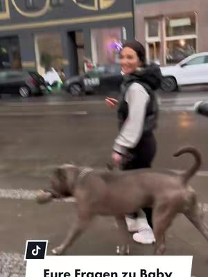 A woman is walking a dog on a wet street, with a cityscape background. The woman is smiling and looking towards the camera. The dog is a large breed, walking in front of the woman.