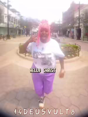 A person with pink hair and a white t-shirt and purple pants is walking towards the camera. Text overlays on the image read ''HELLO CHRIST'' and ''14DEUSVULT8''. The background shows a plaza with buildings and trees.
