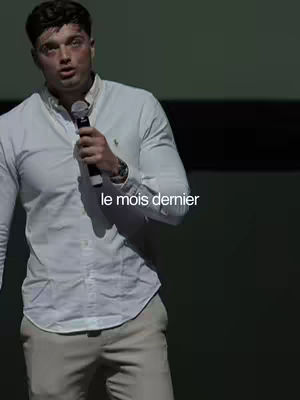 A man in a white shirt and beige pants is holding a microphone and speaking. Text overlayed on the image reads "le mois dernier".
