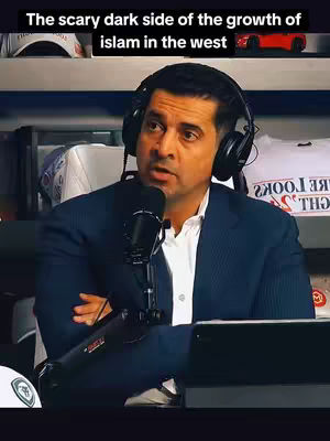 The image shows a man with dark hair wearing a black headset and a dark blue suit jacket over a white shirt. He is speaking into a microphone. The background includes what appears to be a shelf with white shirts and other objects.