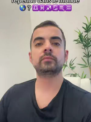 A man with a beard and dark hair is centered in a selfie-style video, framed from the chest up. He wears a dark t-shirt and has a neutral expression. Above his head, text overlays include a globe emoji, question mark, and astrological symbol emojis. The background is a plain white wall with a hint of a green plant visible.
