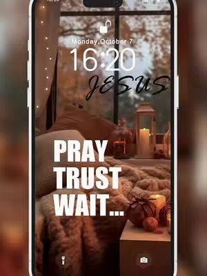 A phone lock screen displaying the time, date, and the word "JESUS" in cursive above the words "PRAY TRUST WAIT..." in bold white text. The background has a warm, inviting aesthetic suggesting a cozy interior scene with a window offering a glimpse of fall foliage. Lit candles and decorative elements add to the serene and inspirational vibe.