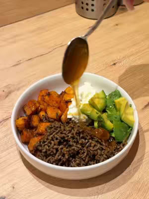 A top-down shot of a white bowl filled with a colorful meal. The bowl contains roasted sweet potatoes, ground beef, cottage cheese, diced avocado, and a drizzle of honey. A metallic spoon is held above the bowl, with honey dripping from it.