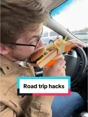 A man is shown driving and about to eat a burger in a car. He is wearing a brown jacket and glasses. Text overlay reads 'Road trip hacks.'