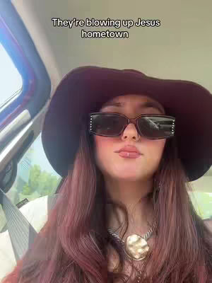 A woman in a car wearing sunglasses and a large hat faces the camera close-up. Text overlayed on the image states, "They're blowing up Jesus hometown."