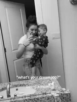 A person holds a phone up taking a mirror selfie of themself and a child in a bathroom. The person is wearing a white t-shirt and jeans, they have their hair up. The child is wearing pajamas, the background is a white door with a sink.