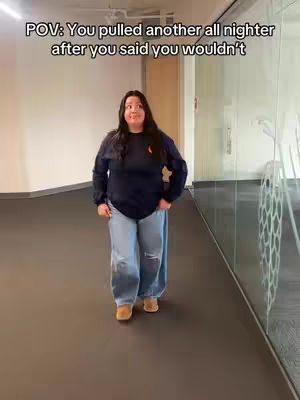 A woman is walking forward in an indoor hallway setting, captured from a full-body perspective. She is wearing a dark blue long-sleeve shirt and light-blue ripped jeans paired with brown shoes. The background shows a pale wall, a hallway with black floor, and a transparent glass panel on her right side