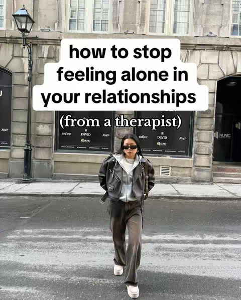 A woman in sunglasses is walking towards the camera on a city street. She wears a black leather jacket, gray hoodie, and wide-legged gray pants. Overhead, a white text overlay reads "how to stop feeling alone in your relationships (from a therapist)". The background features a stone building.