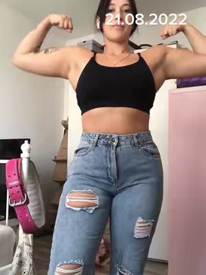 A woman flexes her arms, showcasing her bicep muscles. She wears a black sports bra and ripped jeans. The image has a light color grading, with a pink cabinet visible in the background. The date '21.08.2022' is overlaid in white text.