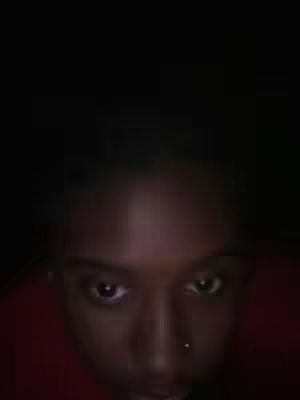 The image features a close-up shot of a person's face. The person is in a dim, low-lit environment, with only their face illuminated. The person has dark skin and their eyes are focused on the viewer. Their hair is out of focus in the background, and they appear to be wearing a red shirt or jacket.