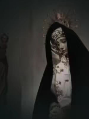 The cover image presents a grayscale shot of a statue or a person in a contemplative pose. Overlaid with shadow, a figure wearing an elaborate head covering and a long black veil that drapes over their face is visible. The figure's facial features are subtly defined, with elements of a halo or decorative crown above the head, set against a dark, textured backdrop. The composition directs focus on the subject's solemn expression.