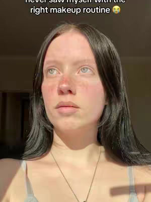 A young woman looks directly at the camera with a slightly concerned expression. Her dark hair is styled with a layered cut. Shadow falls across her face. She wears a delicate necklace and a light-colored top. The background is softly lit.