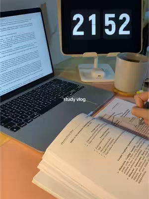 A desk setup with a laptop displaying text, an open book, a digital clock, and a coffee mug. The lighting is warm, suggesting a study environment.