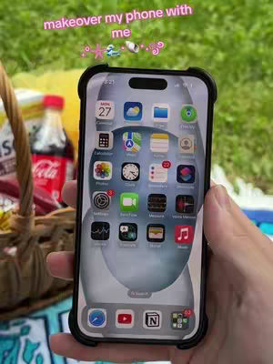 A hand holding a smartphone displays an ocean-themed home screen. The phone is surrounded by a black case, and in the background, there's a picnic area with a drink and food basket. Text floated over the scene.