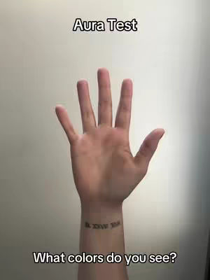 The image shows a hand, palm facing the viewer, against a plain background. The wrist of the hand has a small tattoo with Roman numerals. Above the hand is text that says 'Aura Test', and beneath the hand is text that says 'What colors do you see?' The composition is simple, focusing on the hand and the text. The background is a neutral tone.