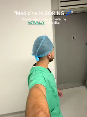 A selfie-style shot from the perspective of a person wearing a medical cap and scrubs. The person has dark hair and a beard, and is looking slightly to the side. The background is a hallway with a door and a sign.