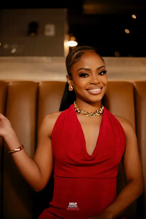 A woman with dark hair and makeup smiles at the camera. She is wearing a red dress and gold jewelry, seated in a booth. The image is well-lit and the background is slightly blurred.