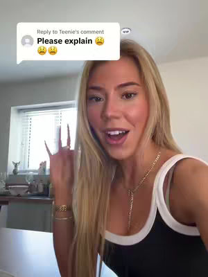 A young woman with long blonde hair is centered in the frame, smiling and gesturing with her hand, with a speech bubble with the words "Please explain". The background is a blurred interior setting.