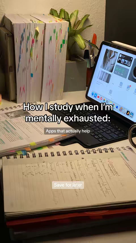 The image shows a desk setup with study materials. There's a large stack of books with colorful sticky notes, a notebook with handwritten notes and equations, and an iPad with a keyboard case displaying the home screen. A green plant adds a natural element. The lighting appears to be natural.