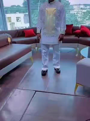 A man in a white suit stands in a modern, well-lit room. The setting includes a large, curving brown leather sofa with red pillows, and large windows.