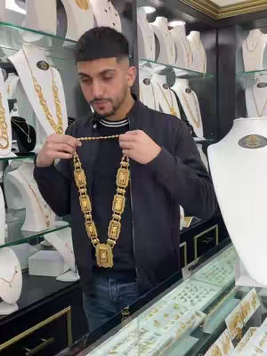 A man stands in a jewelry store holding a gold necklace. The background is filled with display cases of jewelry. The man is wearing a dark jacket and jeans. The necklace is large and ornate.