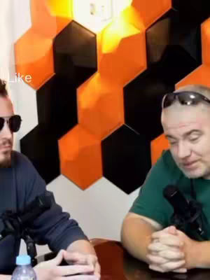 Two men are seated at a table, each holding a microphone. They are positioned in front of a wall decorated with a geometric pattern featuring black and orange hexagonal shapes. The man on the left is wearing sunglasses and a dark shirt, while the man on the right has sunglasses on his head and is wearing a green shirt.