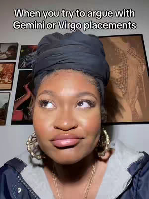 A selfie-style shot of a young woman with a dark head wrap, looking up and to the left with a slightly skeptical expression. The background shows a wall with framed artwork. The lighting appears to be soft, highlighting her features.